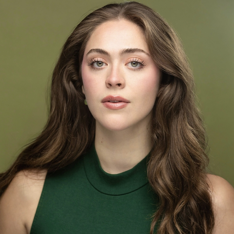 A young woman with long, wavy brown hair and light skin gazes confidently at the camera. Styled by Tanner McIntyre, she wears a sleeveless forest green top, natural makeup with soft pink lips and defined brows, set against a muted olive green background.
