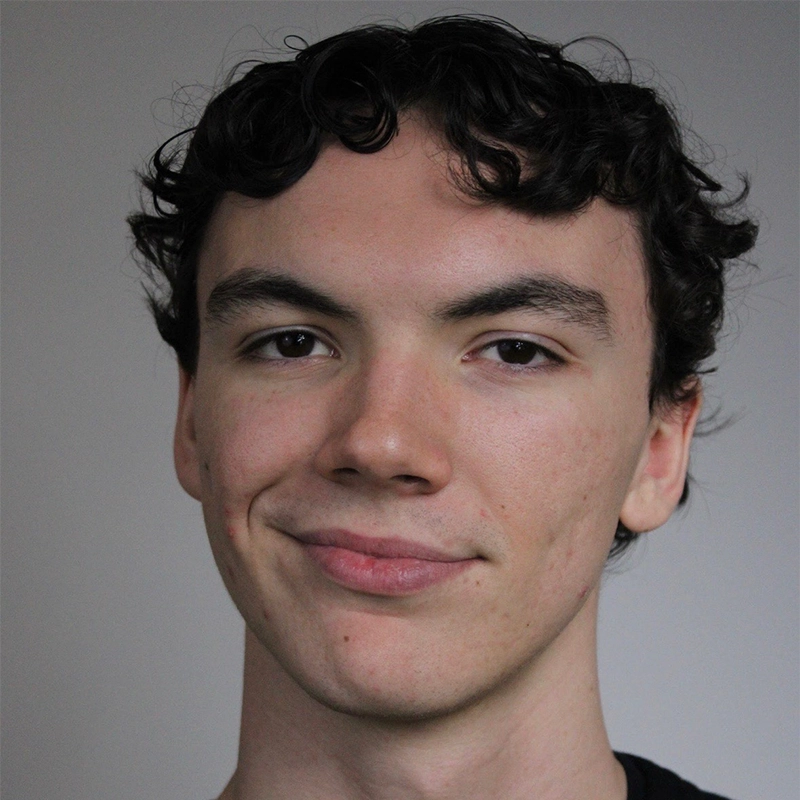 A young person with short, curly dark hair smiles slightly at the camera. Their skin is light, and they wear a black shirt. The plain, neutral background draws attention to Amanda Negrete’s relaxed and confident facial expression.
