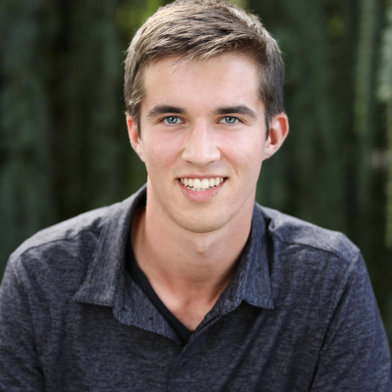 Luca Millard-Kish, a young man with short brown hair and blue eyes, smiles at the camera. He is wearing a dark gray collared shirt over a black T-shirt, standing outdoors against a blurred green background.