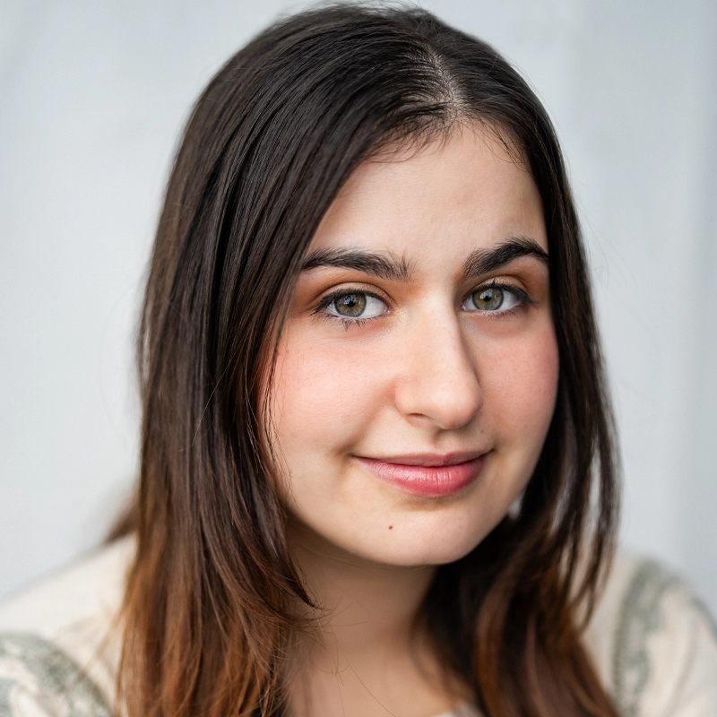 Abigail Mokady, a young woman with long, straight brown hair and hazel eyes, smiles softly at the camera. She wears a light-colored top and is posed against a plain, light background, her expression calm and friendly.
