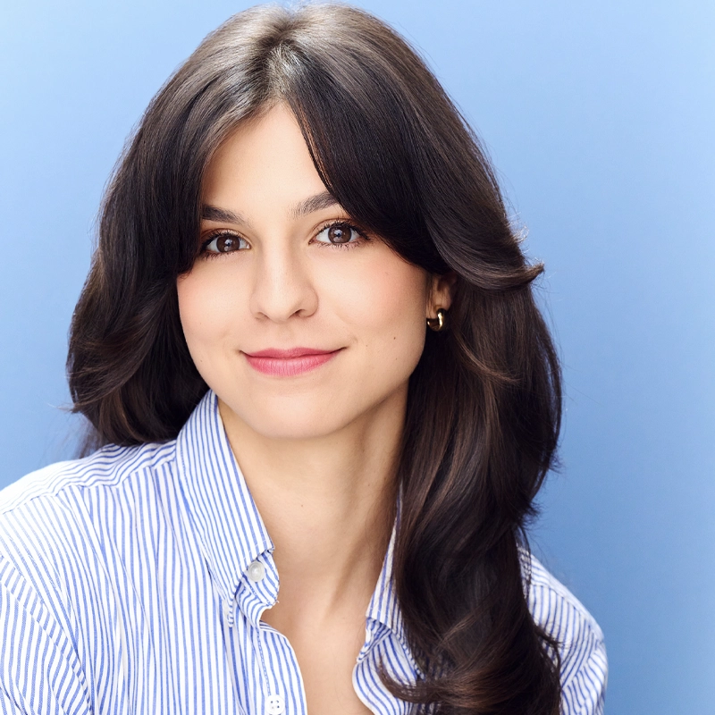 Payton Moledor smiles softly, her long, dark brown hair in loose waves. She wears a blue-and-white striped button-up shirt and small hoop earrings. The solid light blue background adds to the bright, professional headshot.