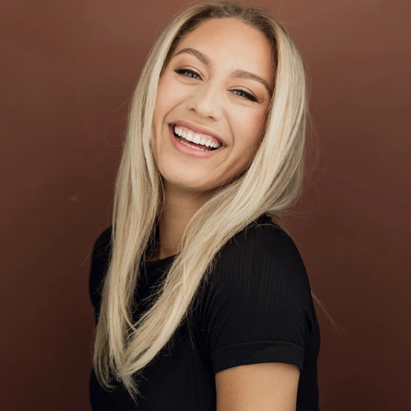 A young woman with long, straight blonde hair and light skin smiles widely, showing her teeth. She wears a black short-sleeve top and faces the camera at an angle, with a brown background behind her.