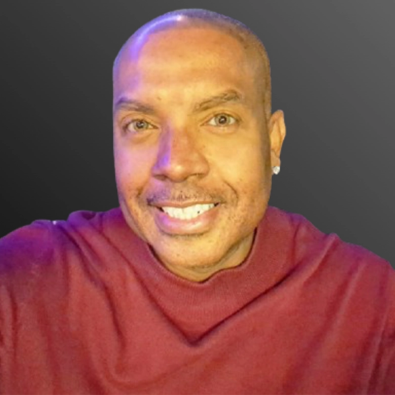 Reginald Moyes, a smiling man with light brown skin, short-cropped hair, and a stud earring, wears a maroon high-collared sweater. The background is a gradient of dark gray as he faces the camera directly under soft lighting.