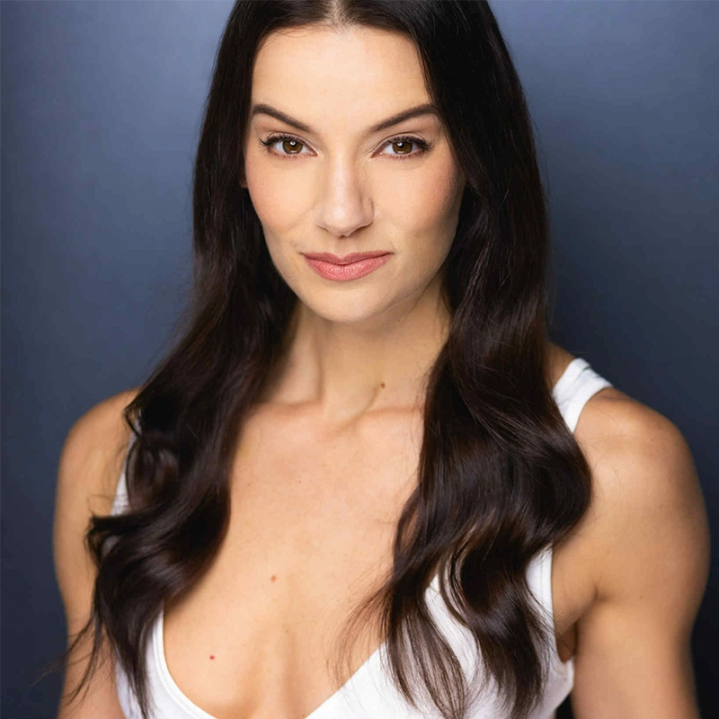 Nicole Benoit, a woman with long, dark wavy hair and light skin, wears a white sleeveless top. She faces forward, smiling softly and posing against a dark blue background, with lighting that highlights her face and hair for a professional headshot look.