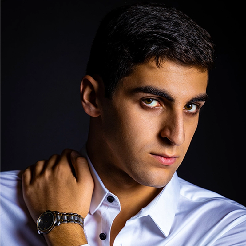 A young man with short dark hair and intense eyes looks at the camera. He wears a white button-up shirt and a metal wristwatch, with one hand resting on his shoulder. The background is dark, highlighting his facial features and expression.