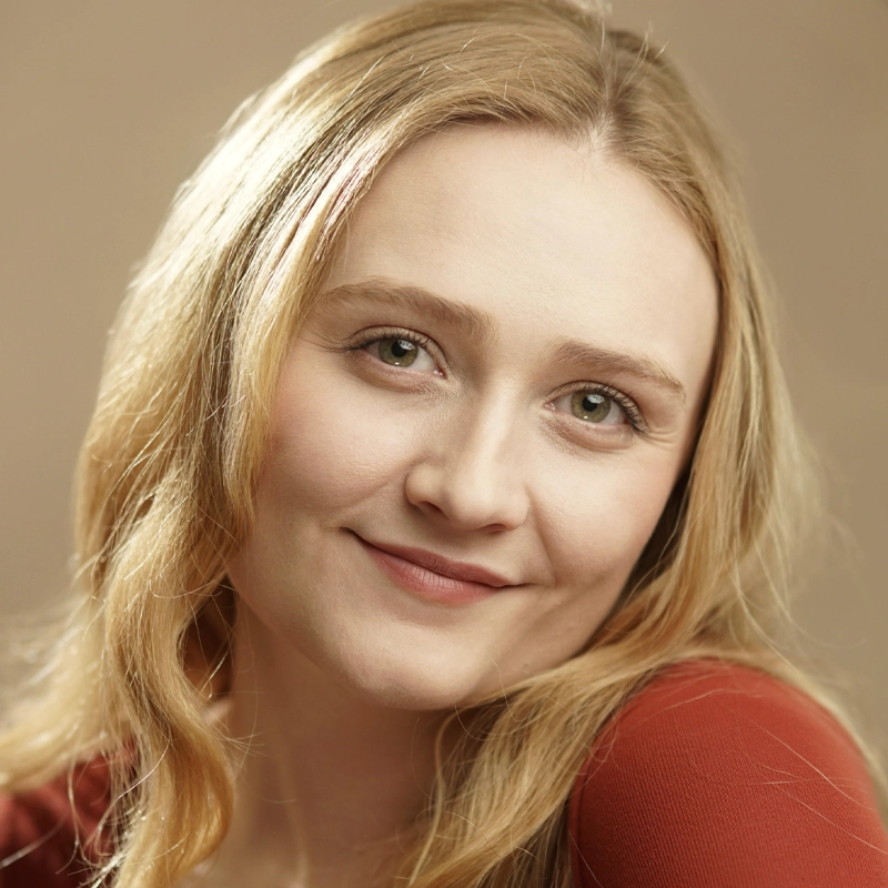 Sarah Beth Palmer, a young woman with long, wavy blonde hair and fair skin, smiles gently in a red top against a soft beige background. Her head is slightly tilted as she looks directly into the camera with a warm, relaxed expression.