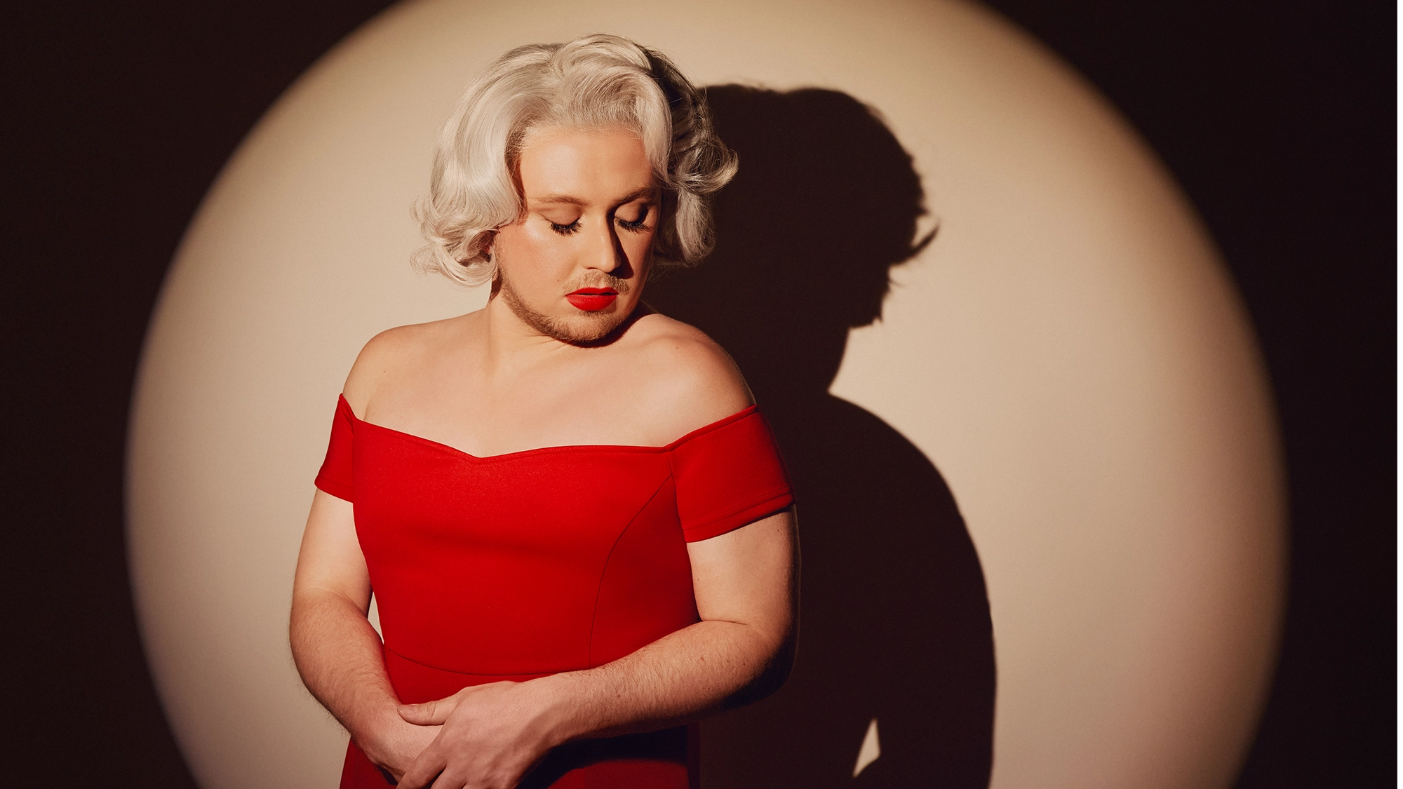 Paloma Diamond site image 25 A person with wavy platinum blonde hair and red lipstick wears an off-the-shoulder red dress. Under a circular spotlight, their hands intertwined, Paloma Diamond gazes down thoughtfully as their shadow is cast on the wall behind them.