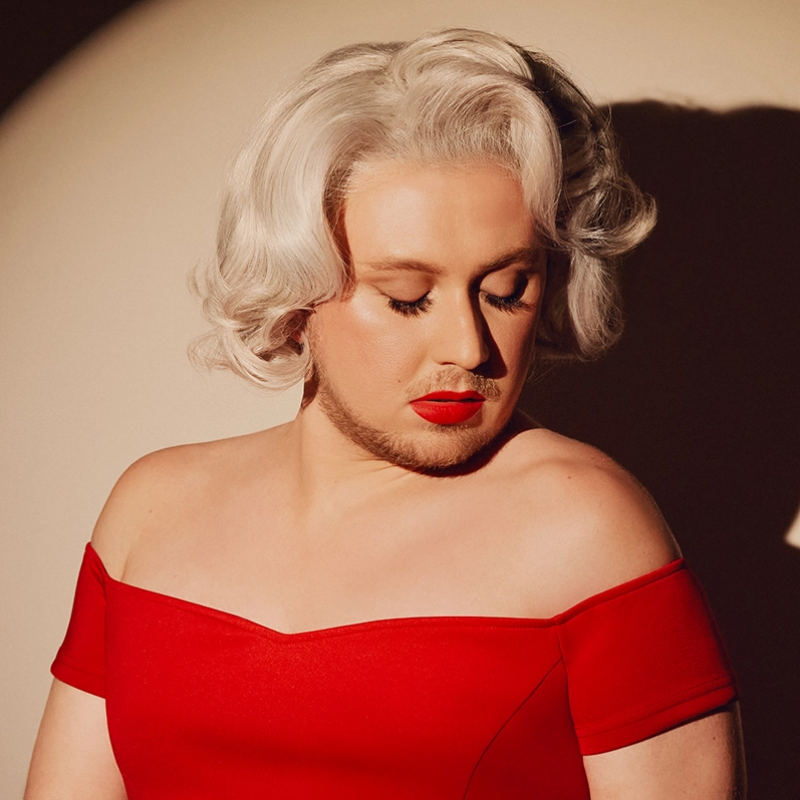 A person with short, wavy platinum blonde hair and red lipstick wears an off-the-shoulder red dress. They look down, eyes closed, as light casts a shadow behind them. Their makeup is dramatic, with contoured cheeks and long eyelashes.