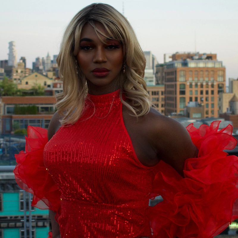A person with long, wavy blonde hair wears a sparkling red halter dress with dramatic ruffled sleeves. They stand outdoors on a rooftop at sunset, with city buildings and greenery visible in the background. Their expression is confident and calm.