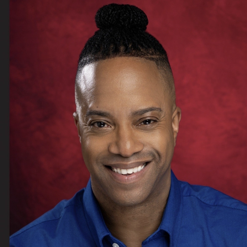 Kwame Michael Remy, a smiling man with medium-dark skin, wears a bright blue collared shirt. His hair is braided into a neat bun atop his head as he poses against a deep red textured background, looking directly at the camera.
