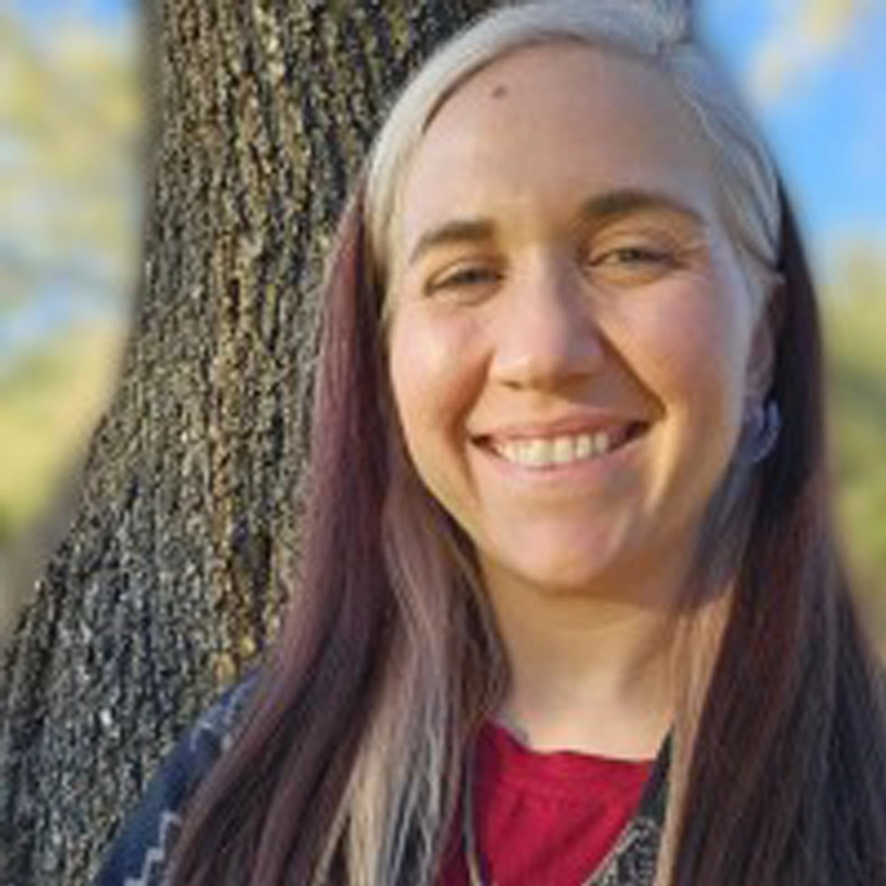 Renee Bandes, with long, straight, brown and white hair, stands outdoors smiling warmly. Wearing a patterned sweater over a red shirt by a tree trunk, she is framed by softly blurred greenery and hints of blue sky in the background.
