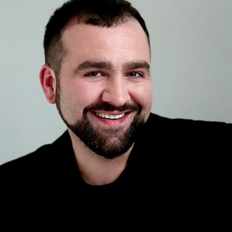 Riad Ymeri Hayden DeWitt, a smiling man with short dark hair and a neatly trimmed beard, wears a black shirt as he poses against a plain, light gray background, facing the camera with a friendly expression and bright eyes.