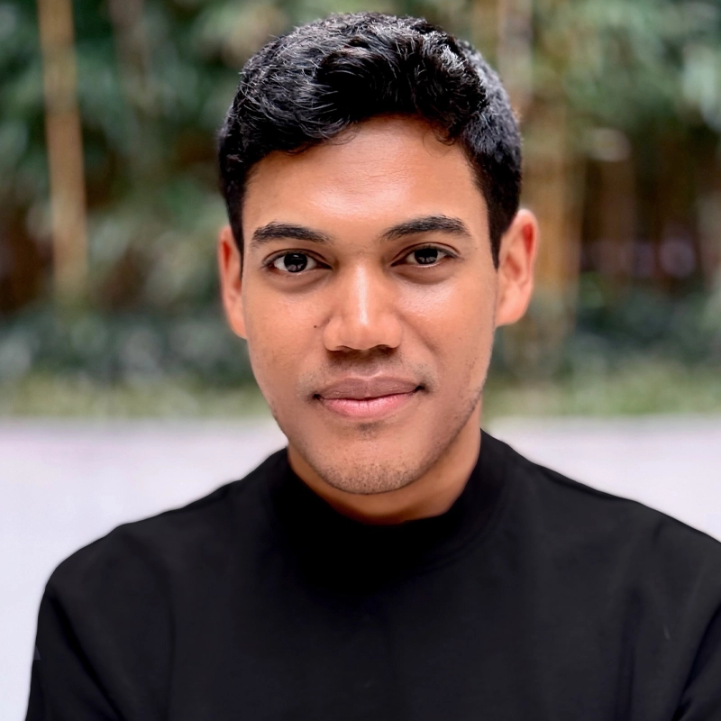 A young man with short, black hair and medium brown skin wears a black turtleneck sweater. He faces the camera with a slight smile. The blurred background features green foliage and natural light.