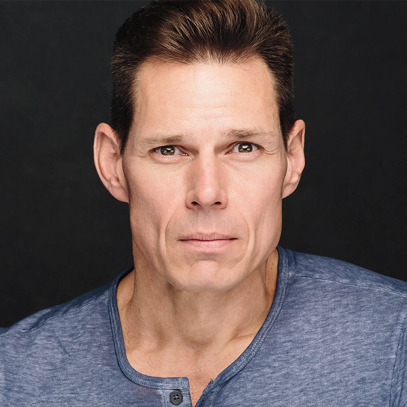 Ryan Williams, a middle-aged man with short brown hair and light skin, looks ahead with a neutral expression. He is wearing a blue button-up shirt with the top button open. The plain, dark background focuses attention on his face.