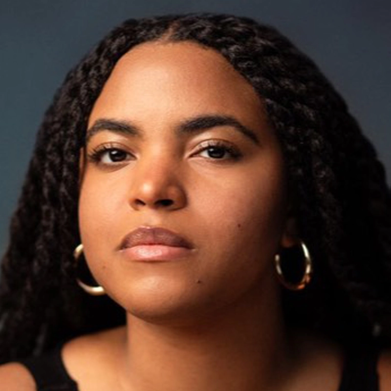 A woman with medium brown skin and long, thick, black braided hair looks directly at the camera. Savannah Lee Birdsong wears large gold hoop earrings and a black top. The dark, blurred background highlights her calm, serious expression.