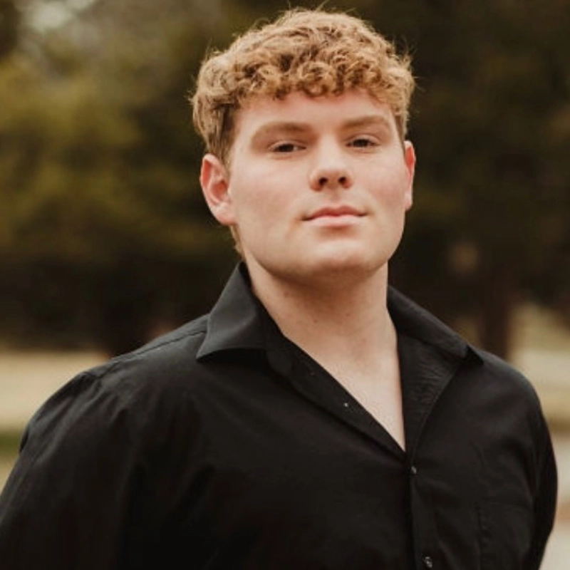 Chris Scanlon, a young man with short, curly light brown hair, stands outdoors looking confidently at the camera. He wears a black button-up shirt, unbuttoned at the collar, with blurred greenery in the background suggesting a park setting.