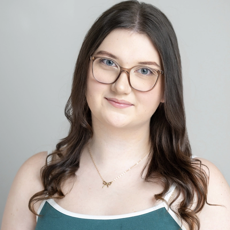 Riley Scotts, a young woman with long, wavy brown hair and fair skin, wears glasses, a green tank top with white straps, and a delicate gold necklace. She stands against a plain light gray background, smiling softly at the camera.