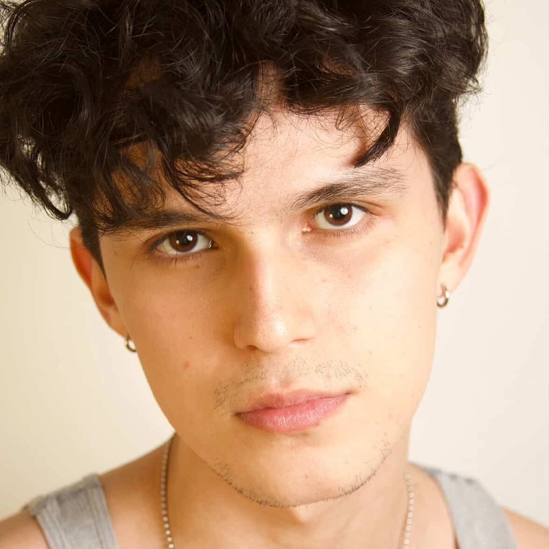 A young person with short, wavy dark hair and brown eyes looks directly at the camera with a neutral expression. They have light skin, light facial hair, and wear small hoop earrings, a gray tank top, and a silver necklace on a plain light background.