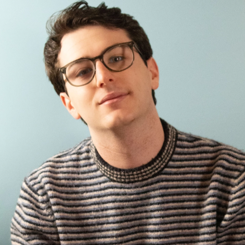 Ethan Shavelson, a person with short, dark hair and glasses, sits against a plain light blue background wearing a striped sweater. Tilting their head slightly, Ethan looks at the camera with a neutral expression.
