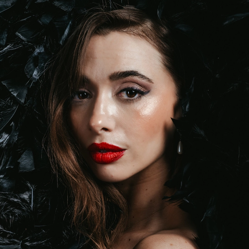 A woman with wavy brown hair and bold red lipstick gazes at the camera. She has shimmery eye makeup, wears a single dangling pearl earring, and is surrounded by black feathers, creating a dramatic and elegant atmosphere.
