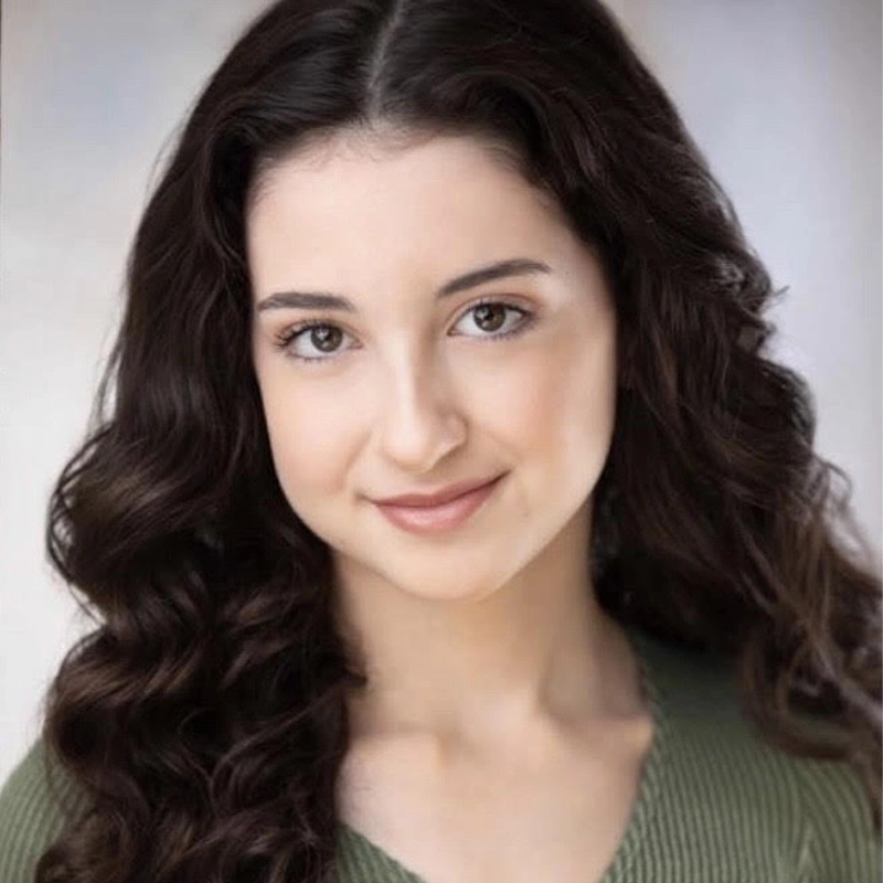 Alyssa Tanti, a young woman with long, dark wavy hair, smiles softly at the camera. She has fair skin, brown eyes, and wears a green ribbed top. The softly blurred background in light tones draws focus to her face.