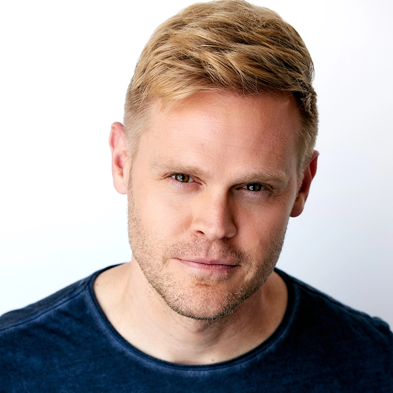 Ryan Williams, a man with short, light blond hair and fair skin, looks into the camera with a neutral expression. He has light stubble and wears a navy blue crew-neck shirt. The background is plain white with bright, even lighting.