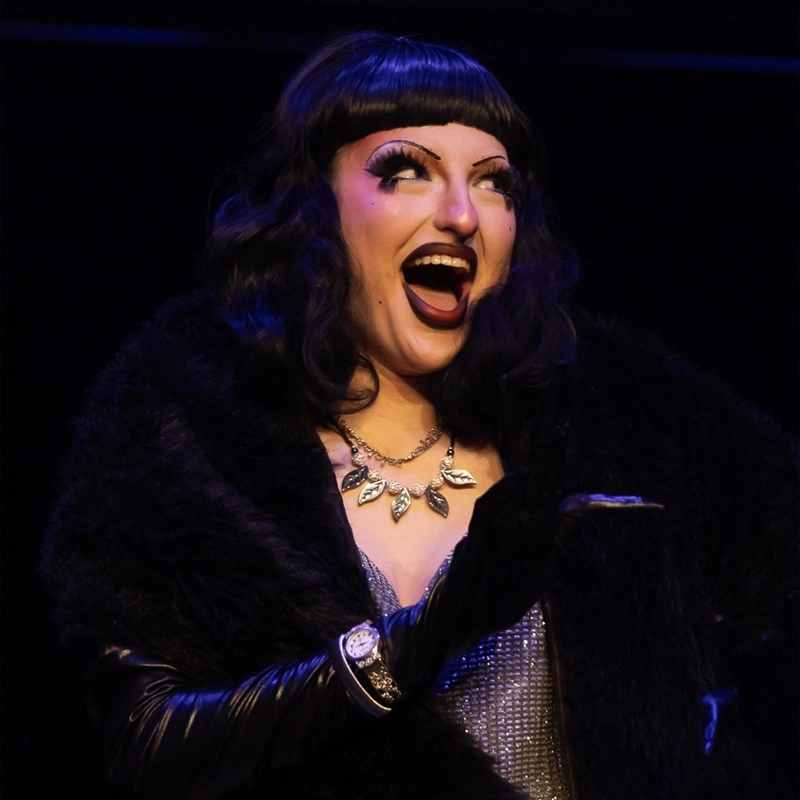 A person with dramatic makeup and a bob haircut smiles widely on stage. Channeling Mae B. Tomorrow, they wear a black fur coat, black gloves, a sparkly necklace, and a silver top, gesturing expressively under stage lighting.