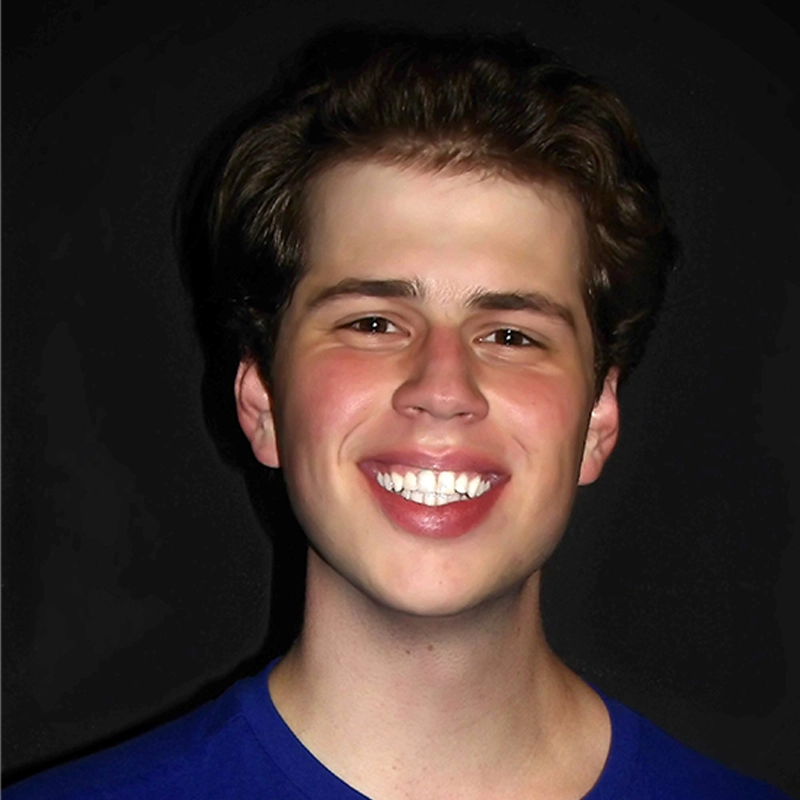 A young person with short, wavy brown hair smiles widely, showing teeth. Warren Mullinnix wears a blue shirt and is posed against a plain, dark background. Their face is well-lit, and their expression is cheerful and friendly.