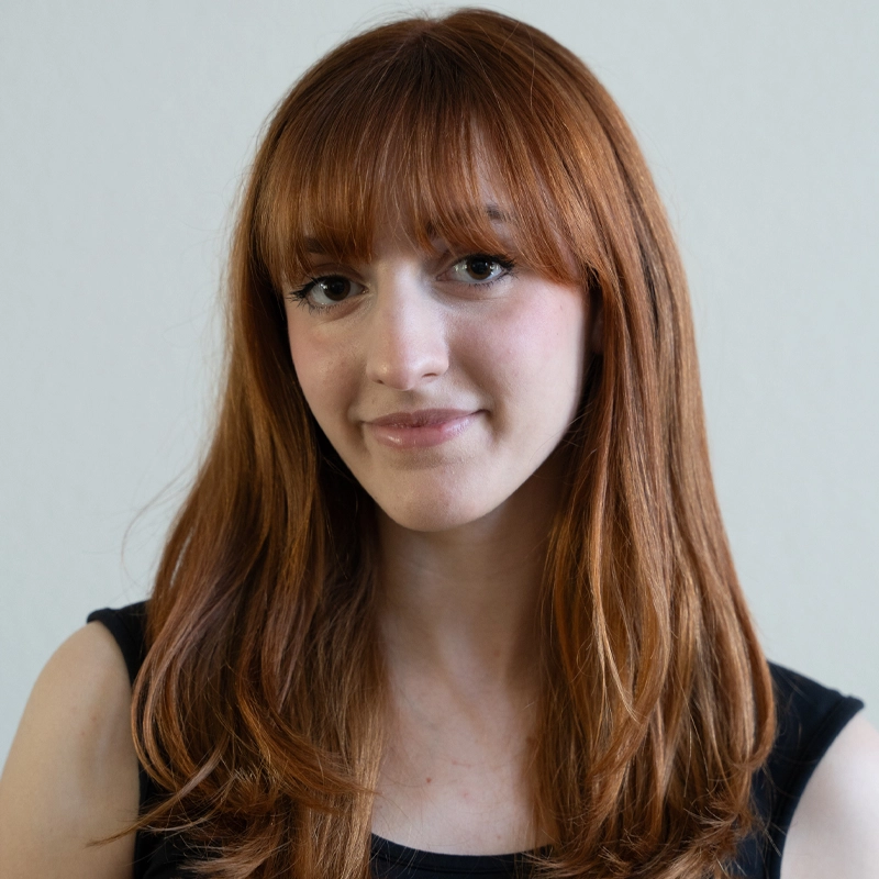Grace Wilkerson, a young woman with straight, long reddish-brown hair and bangs, smiles softly at the camera. She is wearing a sleeveless black top and is posed in front of a plain, light-colored background.