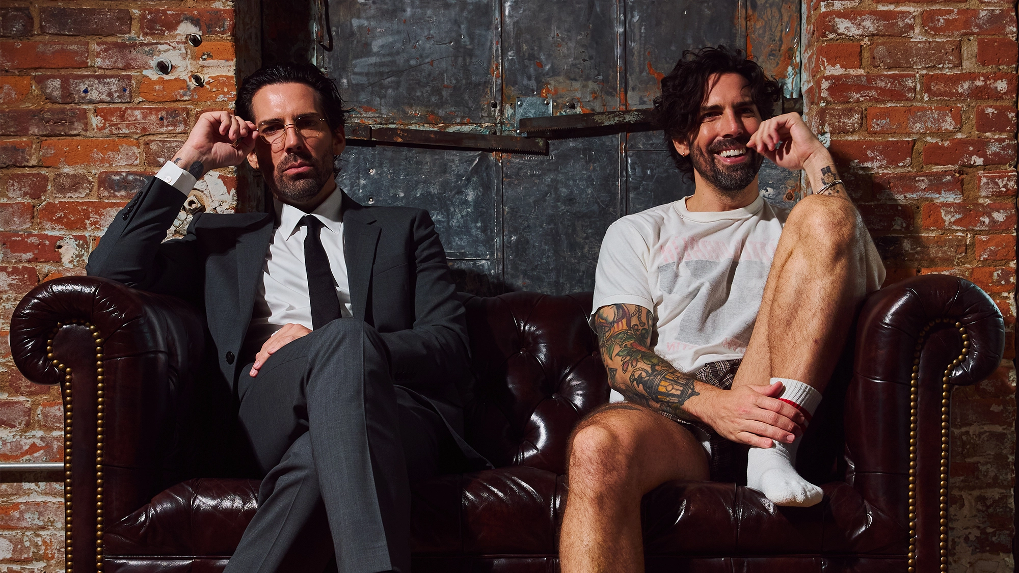 Two men sit on a dark leather couch against a rustic brick wall. Will Kelley Lanier, on the left in a dark suit and glasses, sits formally. The man on the right wears a white t-shirt and shorts, sitting casually with one leg propped up and smiling.