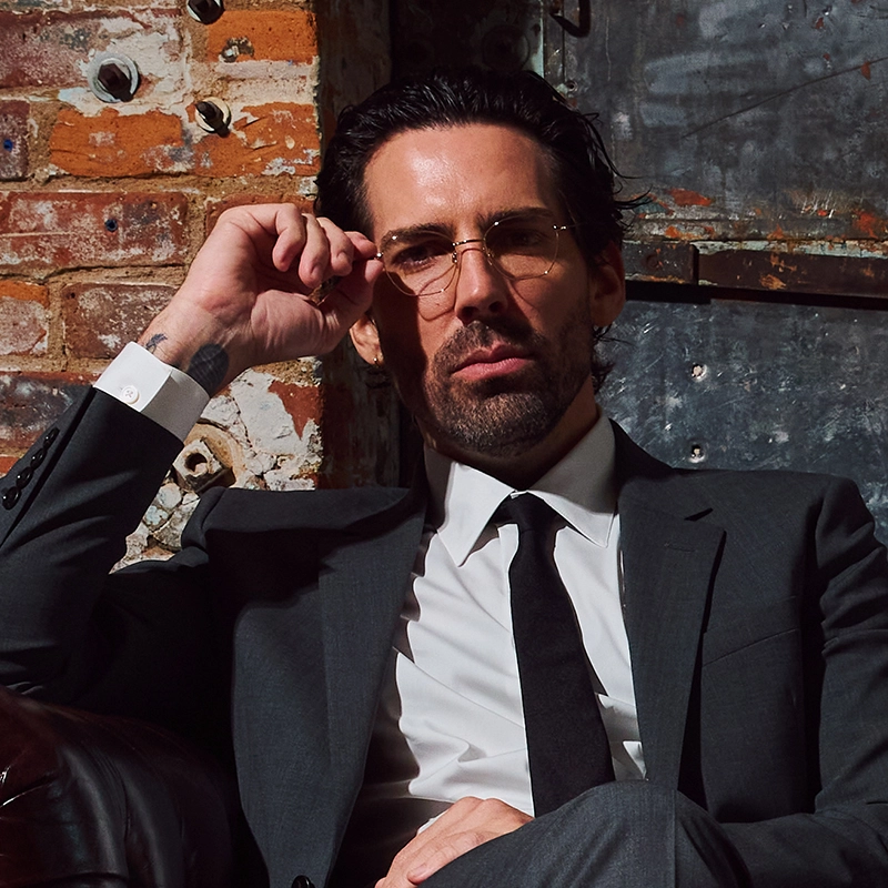 A man with dark slicked-back hair and a trimmed beard, wearing a gray suit, white shirt, and black tie, sits against a weathered brick and metal wall. Will Kelley Lanier gazes at the camera over rimmed glasses, his hand on his face in a confident pose.