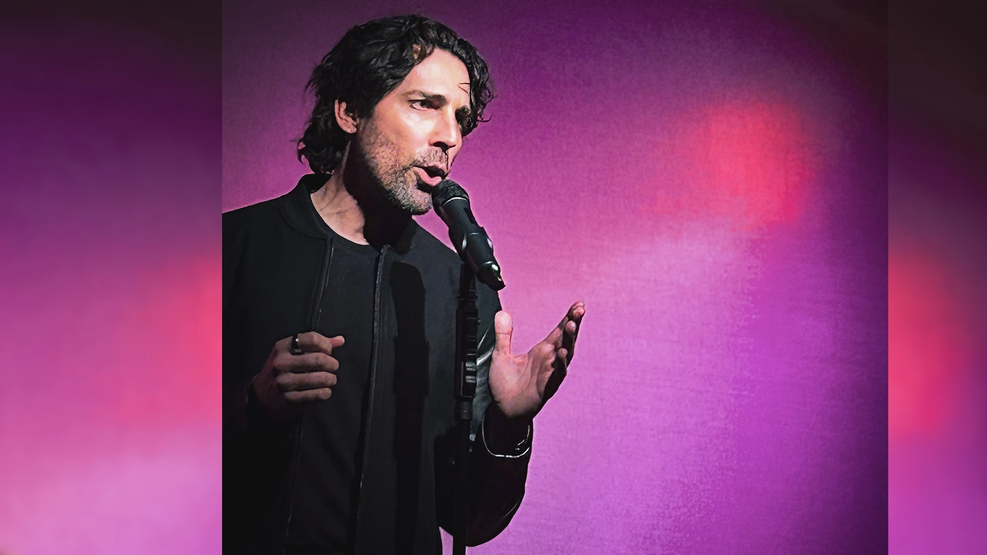 Will Lanier site image 25 A man with dark, wavy hair sings passionately into a microphone. Channeling Year of Yes energy, he wears a black jacket and gestures with one hand as the background glows with a gradient of pink and purple light.
