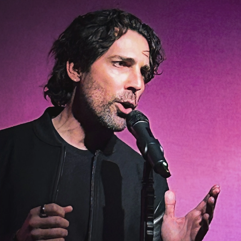 A man with wavy dark hair sings passionately into a microphone, holding one hand up expressively. He wears a black jacket over a dark shirt. The background is a solid, vibrant purple, highlighting his animated facial expression and performance.