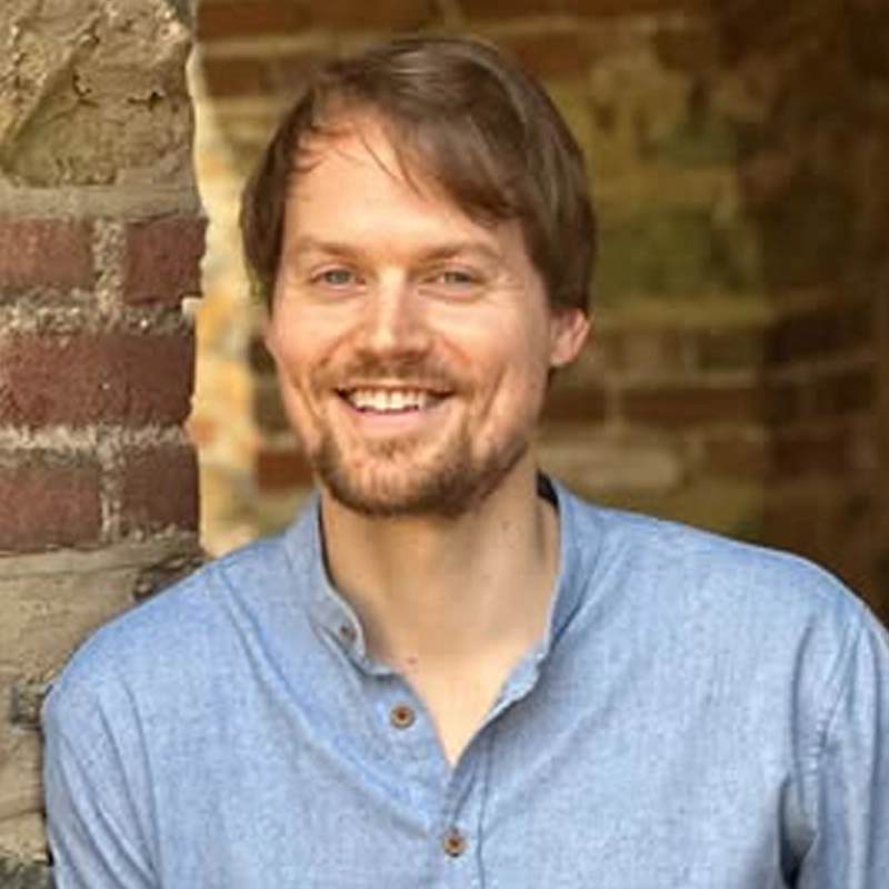 Hayden DeWitt, a smiling man with light brown hair and a short beard, wears a light blue button-up shirt. He stands in front of a textured brick and stone wall, with natural sunlight illuminating his face against the softly blurred background.