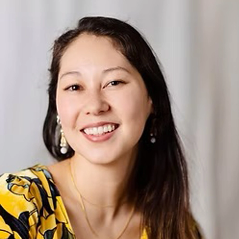 A woman with long dark hair smiles at the camera. She is wearing a yellow patterned top, drop earrings, and a delicate necklace. The background is softly blurred in neutral tones, creating a warm, inviting atmosphere.