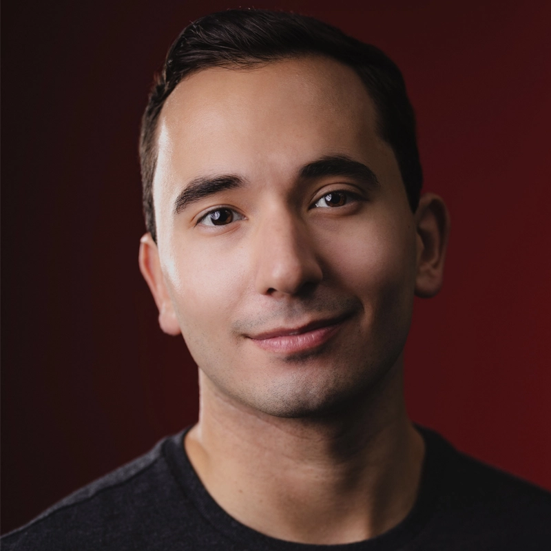 A young man with short dark hair, Caetano de Sá, smiles gently at the camera. He is wearing a dark shirt and is set against a deep red background, with soft lighting highlighting his features and creating a warm, inviting atmosphere.