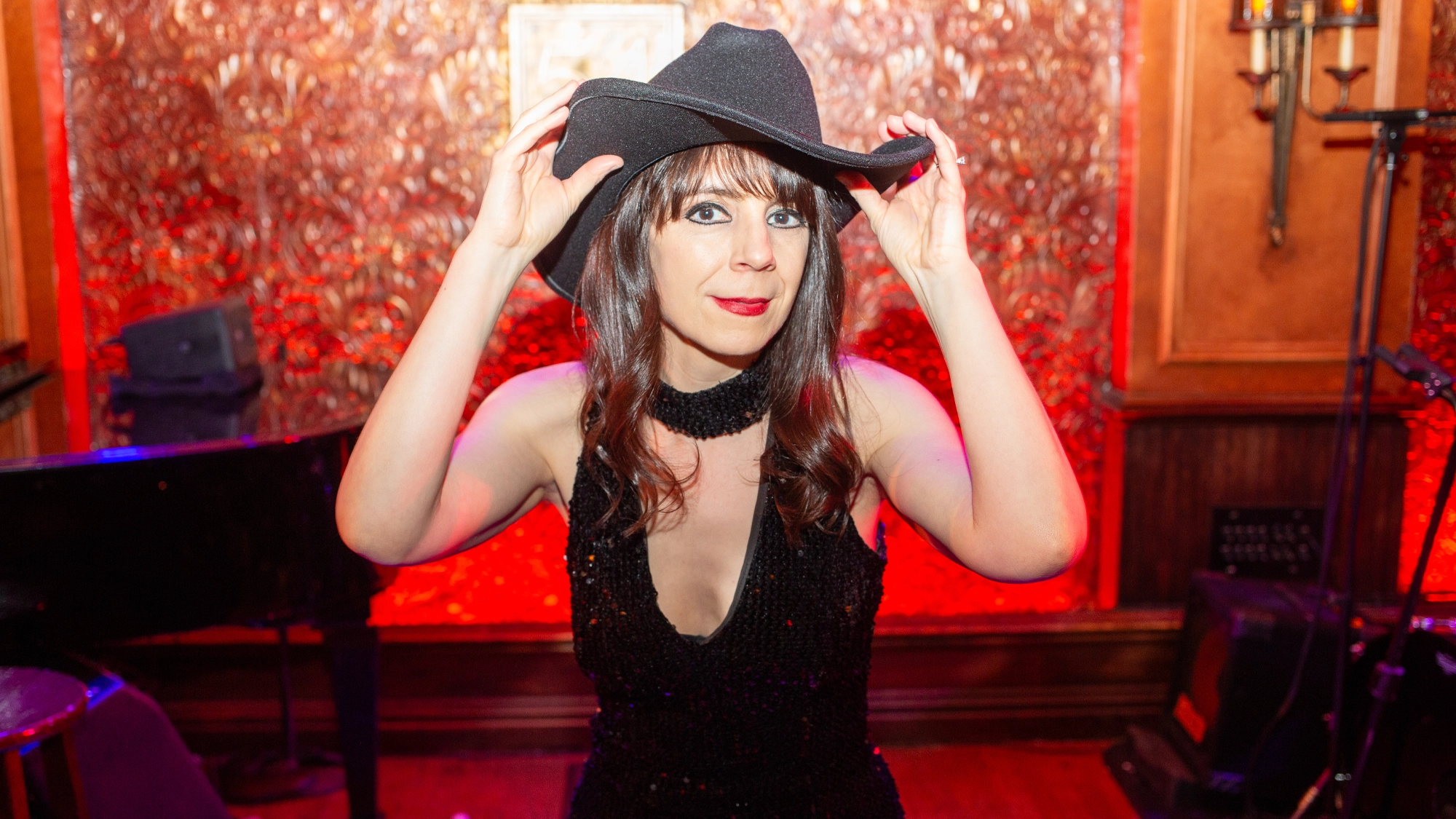 A woman in a sparkly black dress and cowboy hat stands indoors, holding the brim as if ready for a western musical. She has straight brown hair with bangs and red lipstick. Ornate red and gold wallpaper glows in the dim, warm lighting.