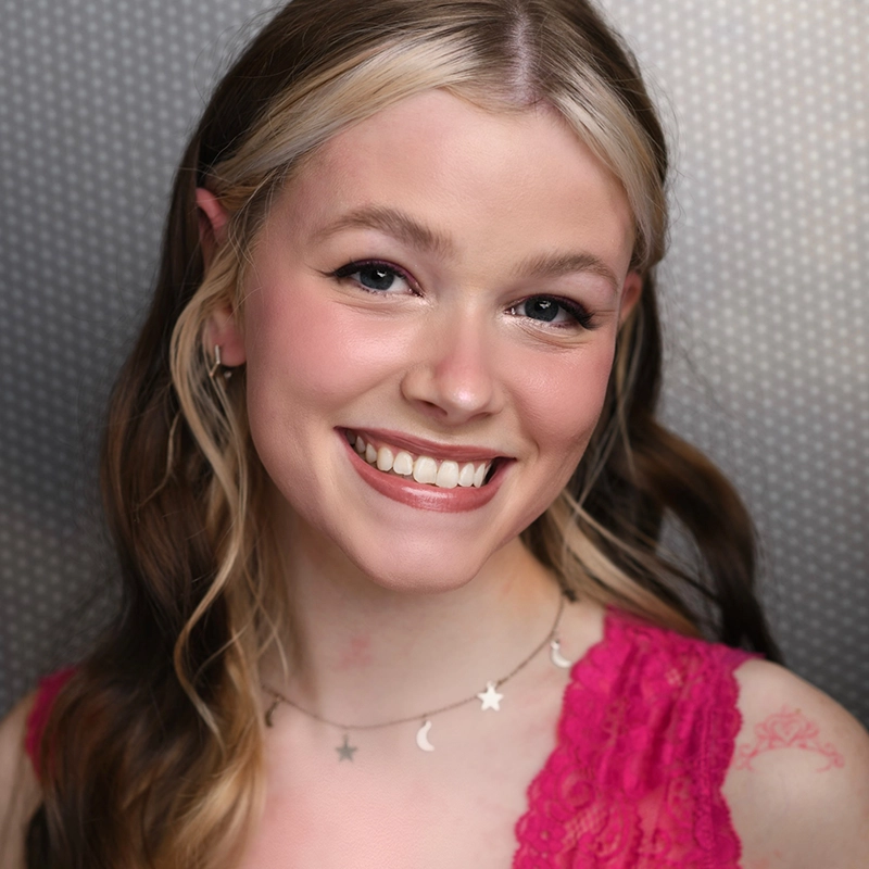 Emma Wallace, a young woman with long, wavy, light brown hair and light skin, smiles at the camera. She wears a pink lace top, star and moon necklace, and has a pink floral tattoo on her shoulder. The background is gray with small white dots.