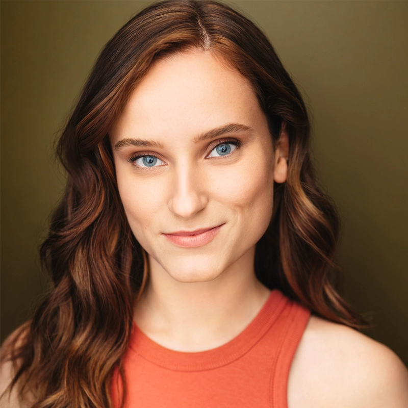 Adriana Ellis, a young woman with wavy brown hair and blue eyes, smiles gently at the camera. She wears a sleeveless orange top against a warm, brownish background. Soft lighting highlights her clear skin and natural makeup.