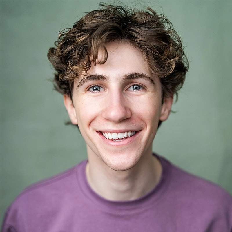 A young man with wavy brown hair and blue eyes smiles at the camera. He is wearing a light purple crew-neck shirt, posed in front of a soft green background—captured by Marielle Utayde with bright, natural lighting that highlights his cheerful expression.