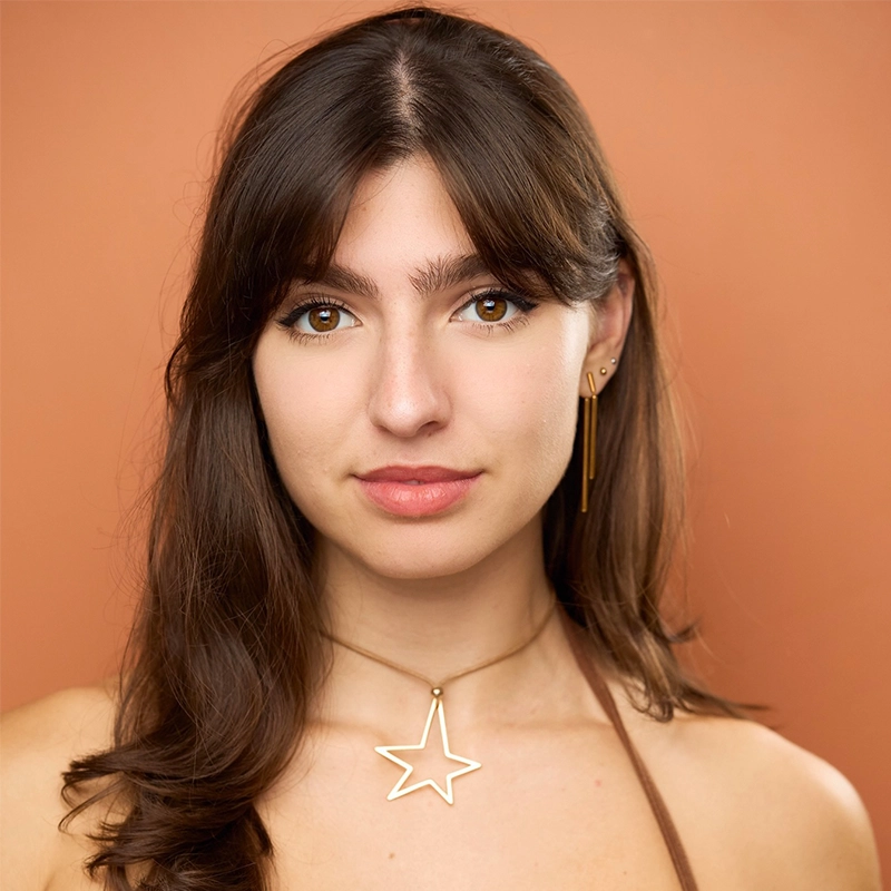 Young woman with long brown hair and bangs poses against a peach background. She wears a gold star necklace and gold earrings by Sergio Guerra, with a natural makeup look and a slight smile. Her bare shoulders suggest a strapless or halter top.