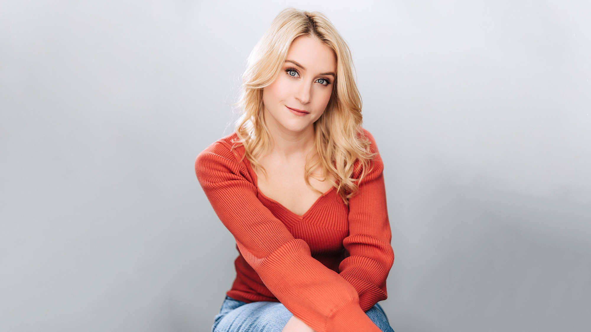 Allison Bailey, a woman with long, wavy blonde hair, sits against a light gray background. She wears a red, long-sleeved sweater and blue jeans, looking at the camera with a gentle smile and relaxed posture.