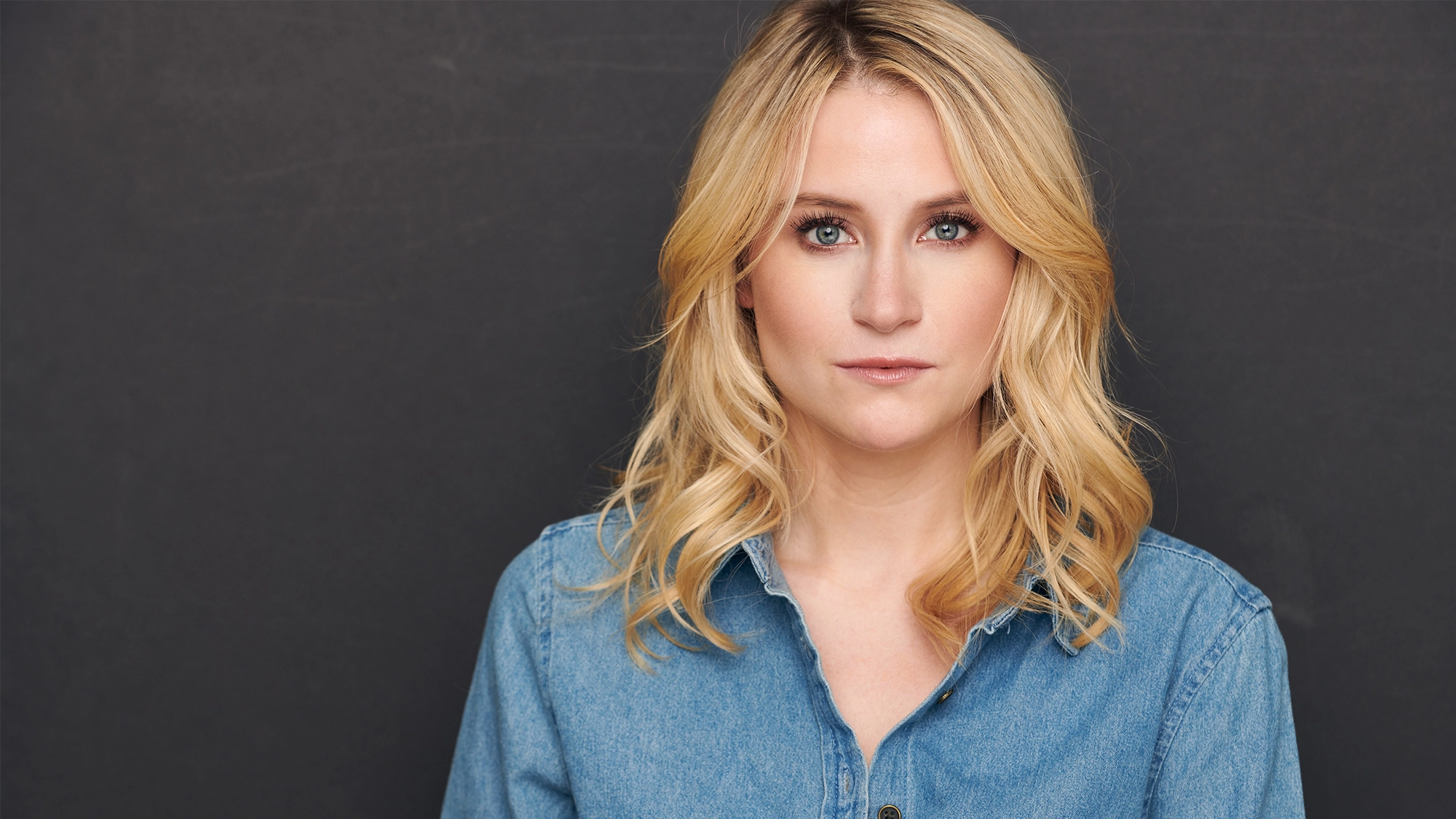 Allison Bailey site image 26 Allison Bailey, a woman with wavy blonde hair and light skin, gazes directly at the camera. She wears a blue denim button-up shirt and stands in front of a dark gray background, her expression calm and neutral with soft, natural makeup.