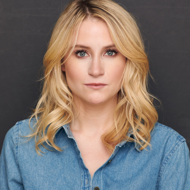 A woman with wavy blonde hair and light skin wears a light blue denim shirt. She looks directly at the camera with a neutral expression. The background is plain and dark, highlighting her face and hair.