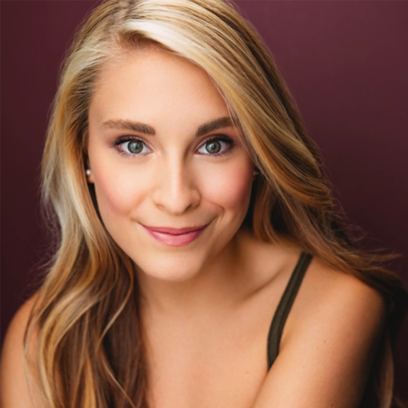 Amanda Harris, a young woman with long blonde hair and blue eyes, smiles gently at the camera. She wears subtle makeup and a dark sleeveless top, set against a solid, deep burgundy background.