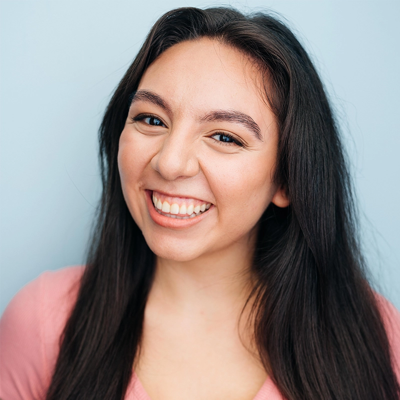 A young woman with long dark hair smiles widely, showing her teeth. She has light skin, dark eyebrows, and is wearing a pink top. The background is a pale blue, giving the image a bright and cheerful feel.