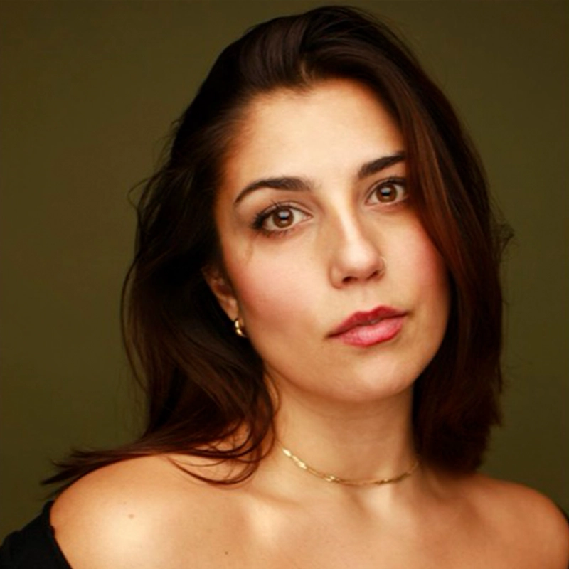 A woman with straight brown hair, parted to the side, gazes softly at the camera. She has light skin, brown eyes, and wears a subtle pink lip color. She is dressed in a black off-the-shoulder top, gold earrings, and a thin gold necklace, with a dark background.