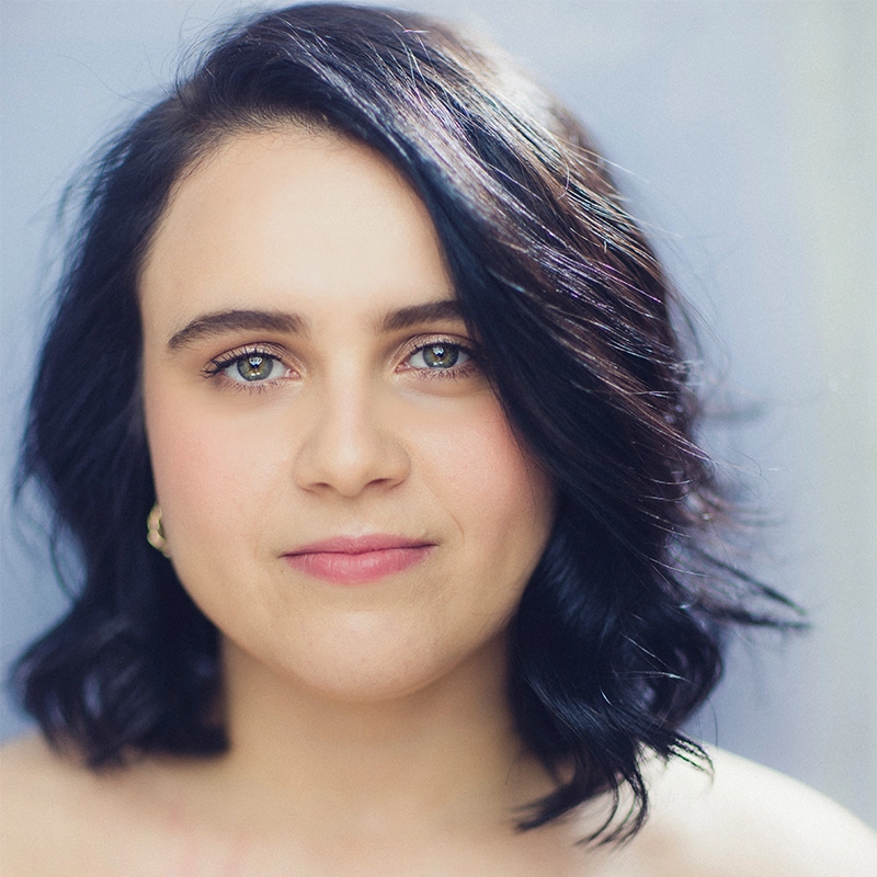 A young woman, Valeria Aceves, with wavy, shoulder-length dark hair and blue-green eyes looks directly at the camera. She has fair skin, subtle makeup, and wears small gold hoop earrings. The background is softly blurred in light gray and blue.