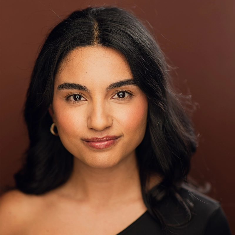 Jaeden Riley Juarez, a young woman with wavy black hair and medium skin tone, looks directly at the camera, smiling softly. She wears a one-shoulder black top and gold hoop earrings against a solid warm brown backdrop for a polished look.