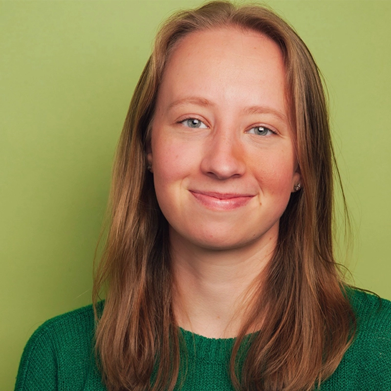 A young woman with fair skin and straight, light brown hair smiles gently at the camera. Analise Levesque wears a green knit sweater and small earrings. The background is a plain, light green color. The image is well-lit and evenly focused.
